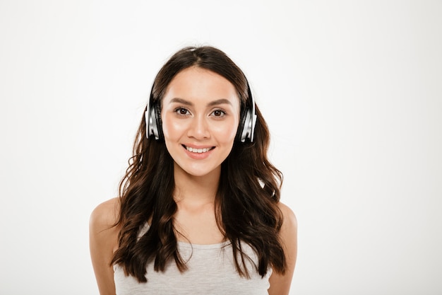 Smiling brunette woman in headphones listening music and looking at the camera over gray