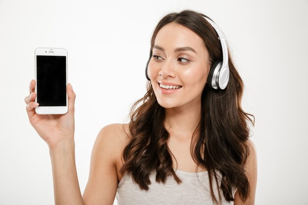 Smiling brunette woman in headphones showing blank smartphone screen and looking at him over gray