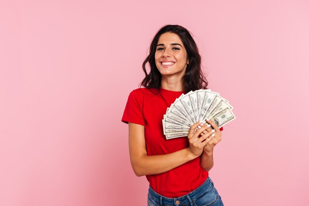 Улыбаясь брюнетка женщина держит в руках деньги и смотрит в камеру над розовым