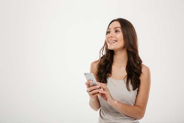 Smiling brunette woman holding smartphone and looking away over gray
