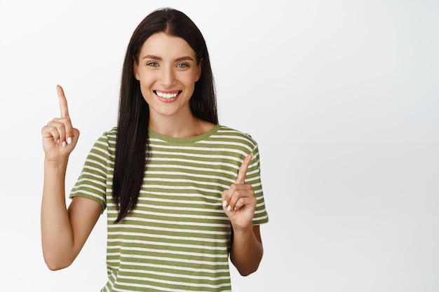 Smiling brunette woman pointing fingers at two ways showing variants standing in tshirt over white background Advertisement concept