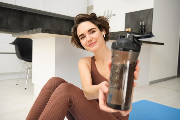Smiling brunette woman sits on yoga fitness mat gives you water bottle to drink after workout does p