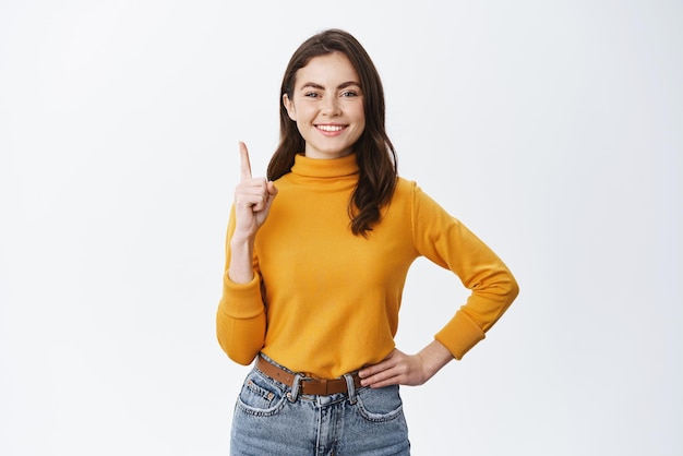 白い背景の上に立っているコピースペースで示す上に広告を示す幸せな女性モデルを指を上に向けて黄色のセーターで笑顔のブルネットの女性
