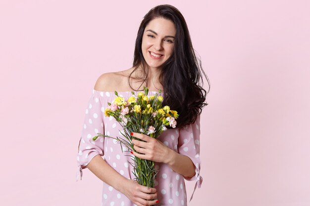 Smiling brunette young European woman has toothy smile, holds bouquet of flowers, wears dress, isolated on pink, rejoices good day, has red manicure. Spring and women concept.