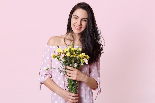 笑顔のブルネットの若いヨーロッパの女性は歯を見せる笑顔があり、花の花束を保持し、ピンクに分離されたドレスを着て、良い一日を喜ぶ、赤いマニキュアをしています。春と女性のコンセプトです。