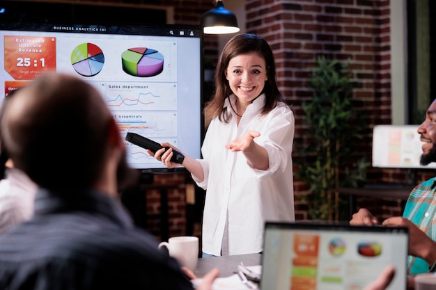 Smiling business employee standing in front of tv screen with sales statistics talking with man with laptop sitting in late night meeting. Woman presenting pie charts and analytics to mixed team.
