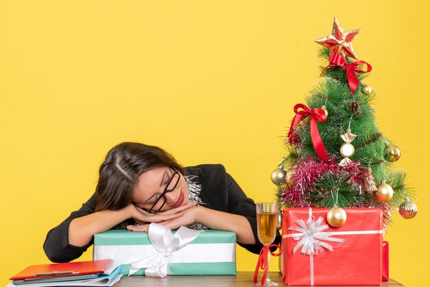 Smiling business lady in suit with glasses sleeping on her gift and sitting at a table with a xsmas tree on it in the office