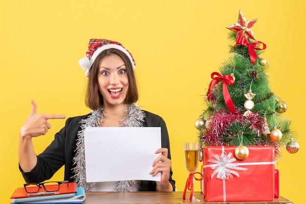 Smiling business lady in suit with santa claus hat and new year decorations working alone pointing documents and sitting at a table with a xsmas tree on it in the office