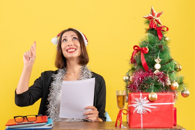 サンタクロースの帽子と新年の装飾とスーツを着て笑顔のビジネスレディが上を指して、オフィスでクリスマスツリーのあるテーブルに座って一人で働いています