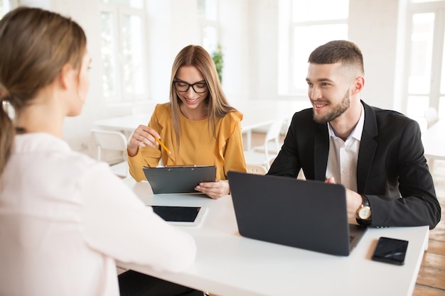 Smiling business man with laptop and business woman in eyeglasses with folder in hand while happily talking with applicant about work Young joyful employers spending job interview in modern office