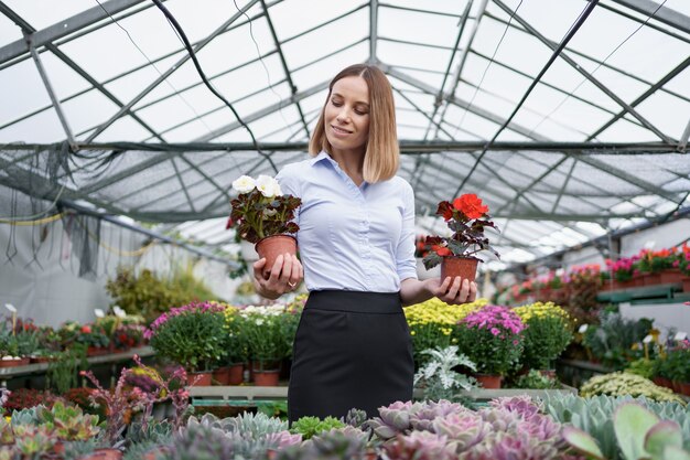 Smiling business owner in her nursery standing holding in hands two pots with red and white flowers in the greenhouse