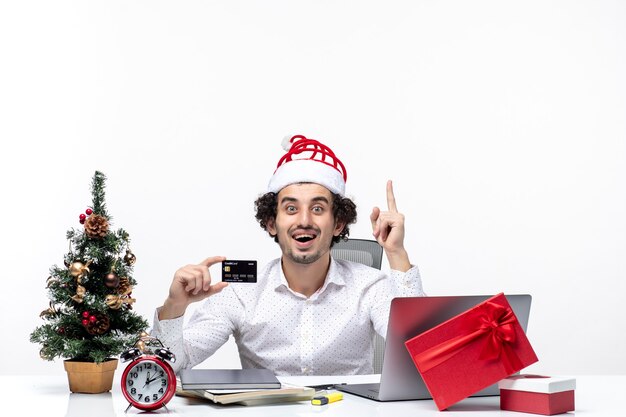 サンタクロースの帽子をかぶって笑顔のビジネスパーソンと白い背景の上のオフィスで彼の銀行カードを指している