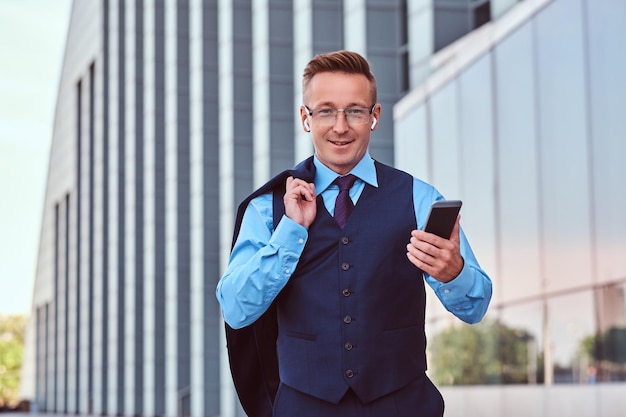 Smiling businessman dressed in an elegant suit holds smartphone and jacket on the shoulder while standing against cityscape background.