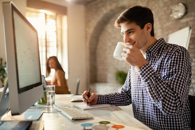 メモを取り、オフィスでデスクトップPCを使用しながらコーヒーを飲みながら笑顔のビジネスマン