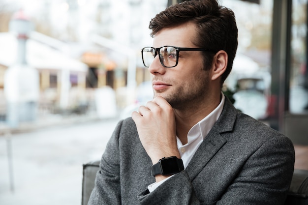 Smiling businessman in eyeglasses sitting by the table in cafe and looking at window