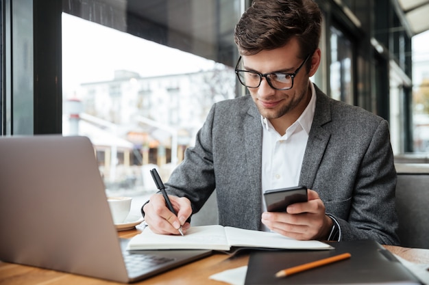 Улыбающийся бизнесмен в очках, сидя за столом в кафе с ноутбуком при использовании смартфона и что-то писать