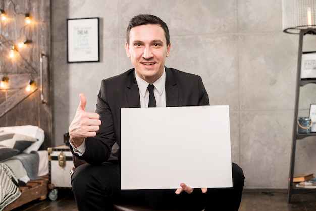 Smiling businessman holding blank paper and gesturing thumb up 