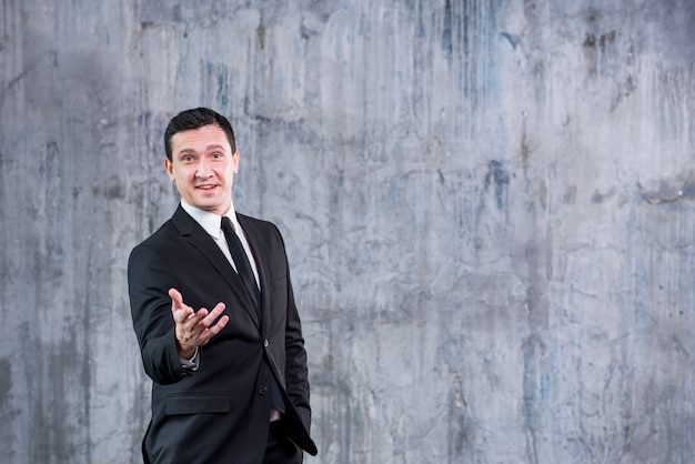 Smiling businessman pointing at camera while standing against grey wall