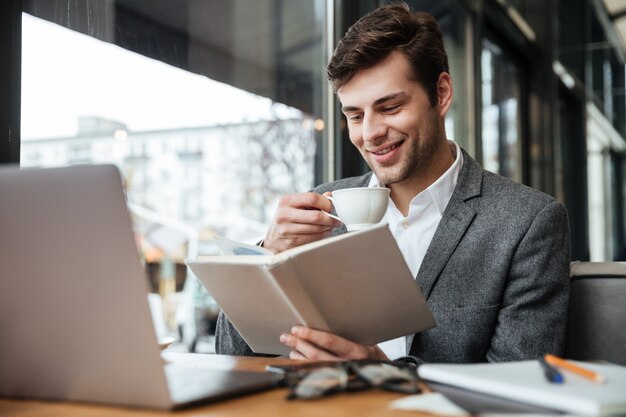 Smiling businessman sitting by the table in cafe with laptop computer while reading book and drinking coffee