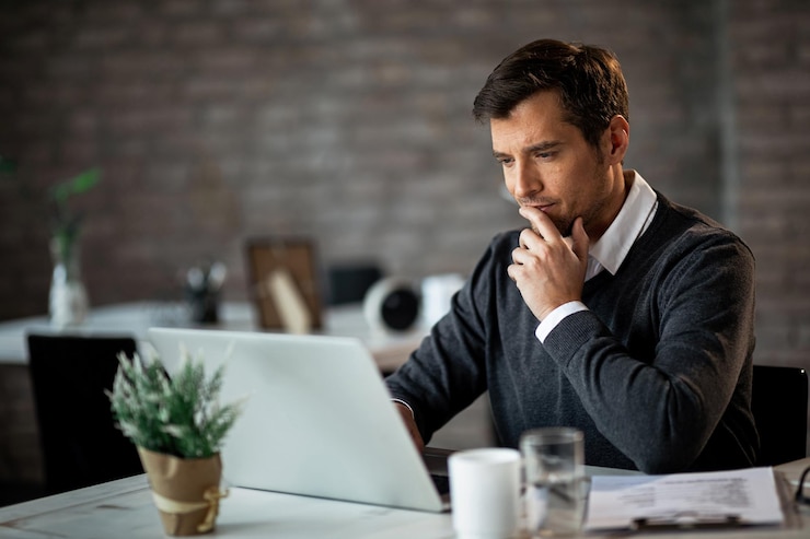 Free Photo smiling businessman using laptop and contemplating while working at his desk in the office