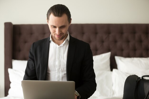 Smiling businessman working on laptop in bedroom.
