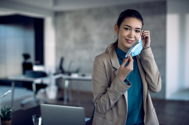 Smiling businesswoman putting on protective face mask in the office and looking at camera