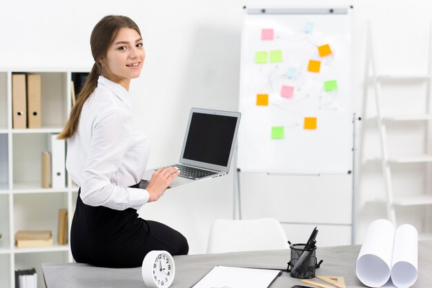 Smiling businesswoman sitting at the edge of table holding digital tablet in hand looking at camera