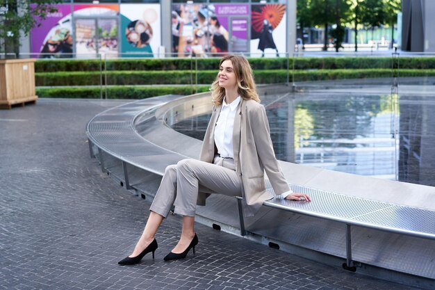 Улыбающаяся деловая женщина сидит возле фонтана и ждет кого-то на улице
