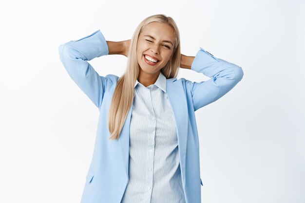 Smiling businesswoman in suit standing relaxed winking and holding hands behind head in resting pose white background