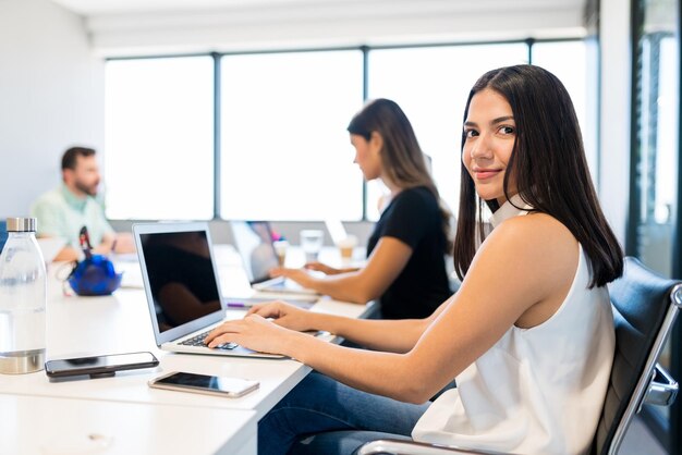 Smiling businesswoman working on laptop while sitting with colleagues at desk in office