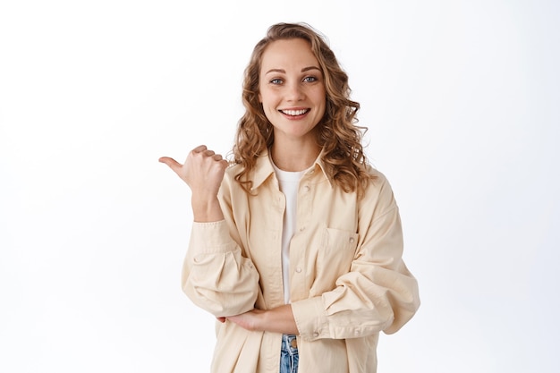 Smiling candid girl with blond curly hair, pointing thumb at left copyspace, showing advertisement, giving recommendation, standing over white wall