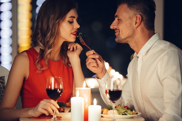 Smiling careful man feed his pretty woman friend while have romantic dinner at home