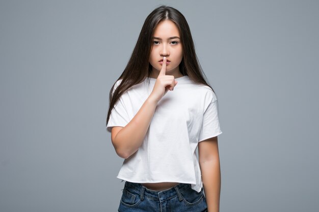 Smiling casual asian woman showing silence gesture with finger over her lips over gray background