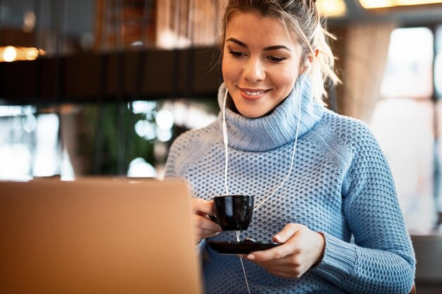 Улыбающаяся случайная деловая женщина просматривает сеть на компьютере и пьет кофе, слушая музыку через наушники в кафе