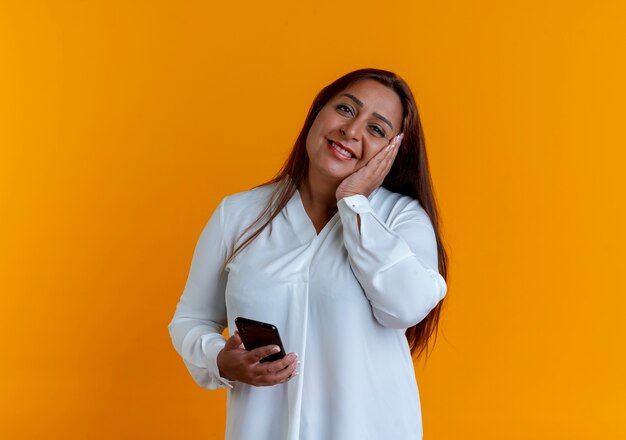 Smiling casual caucasian middle-aged woman holding phone and putting hand on cheek isolated on yellow wall
