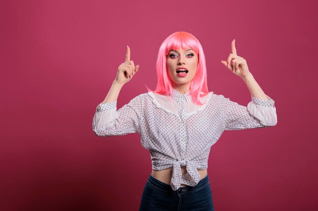 Smiling casual lady pointing up with index fingers in studio, posing with raised arms to indicate advertisement upwards. Indicating and recommending directions with hands in front of camera.