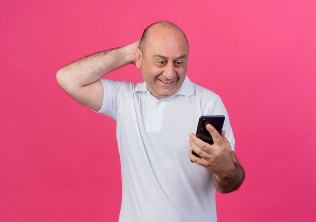 Smiling casual mature businessman holding and looking at mobile phone keeping hand behind head isolated on pink background with copy space