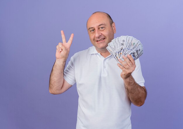 Smiling casual mature businessman holding money and doing peace sign isolated on purple background with copy space