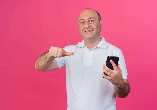 Smiling casual mature businessman holding and pointing at mobile phone isolated on pink background with copy space