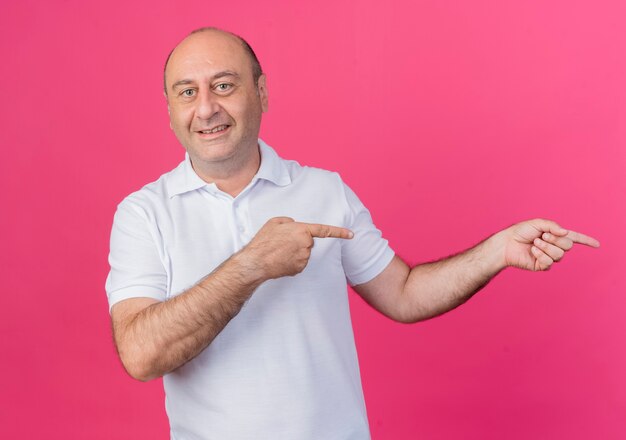 Smiling casual mature businessman looking at camera pointing at side isolated on pink background