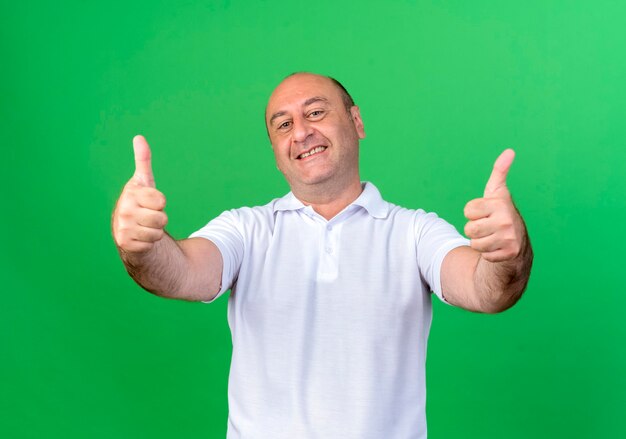 Улыбающийся случайный зрелый мужчина его большие пальцы руки вверх изолирован на зеленой стене