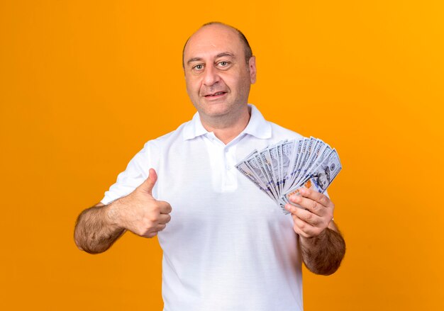 Smiling casual mature man holding cash and his thumb up isolated on yellow wall