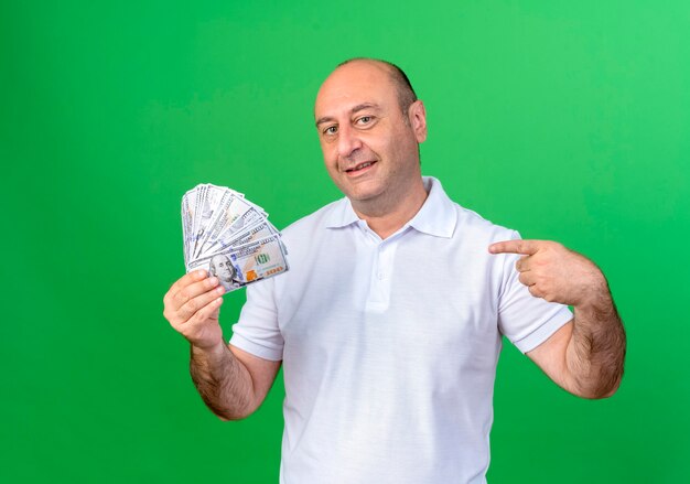 Smiling casual mature man holding and points at cash isolated on green wall