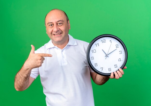 Smiling casual mature man holding and points at wall clock isolated on green wall
