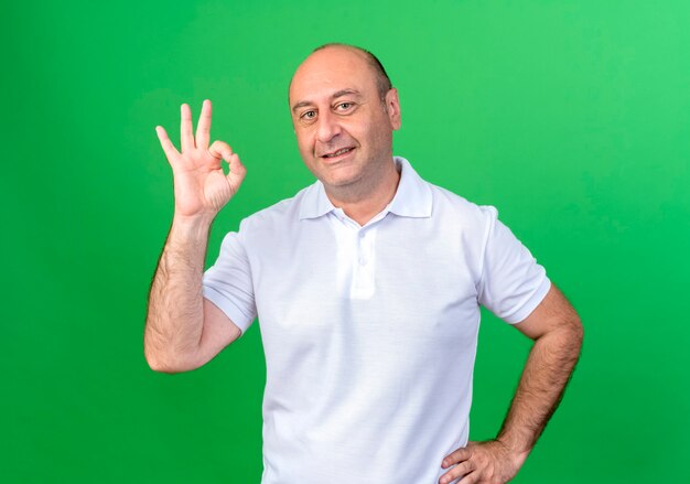 Smiling casual mature man showing okey gesture and putting hand on hip isolated on green wall