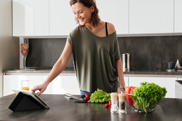 Улыбаясь случайный женщина, стоящая возле стола с овощами на кухне и с помощью компьютера tablt