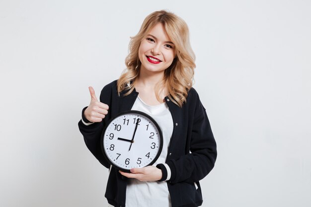 Smiling casual young woman teenager holding clock and showing thumb up