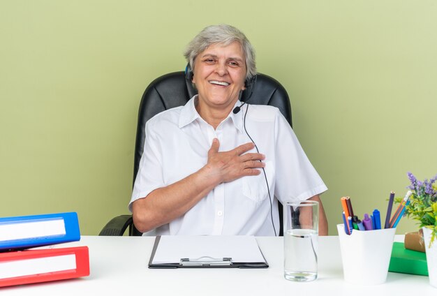 Smiling caucasian female call center operator on headphones sitting at desk with office tools putting hand on her chest isolated on green wall