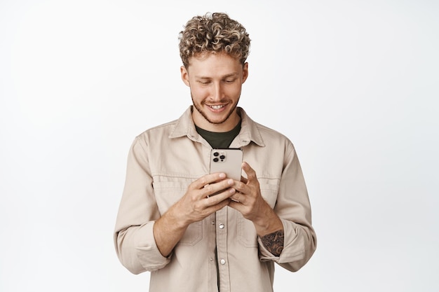 스마트폰을 사용하여 웃는 백인 남자 흰색 배경 위에 서서 휴대전화로 채팅하는 잘생긴 남자