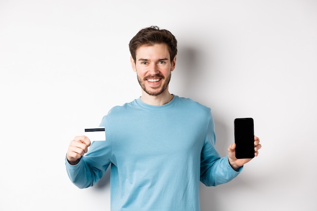 Smiling caucasian man showing plastic credit card with mobile phone screen. Guy recommending online banking app, standing on white background.